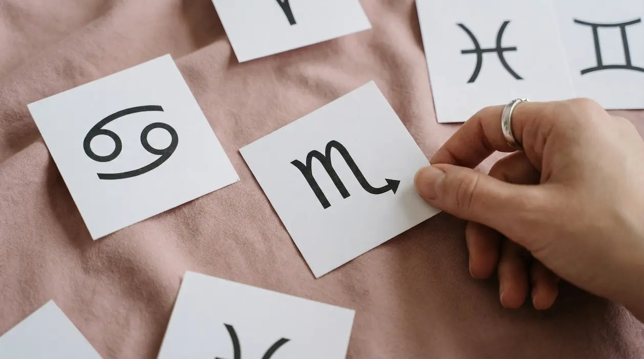 Mano che tiene un cartoncino con il simbolo dello Scorpione tra altri segni zodiacali su un tessuto rosa